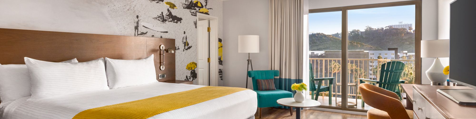 A modern hotel room with a large bed, vibrant accent decor, a seating area, and a balcony with a scenic view.
