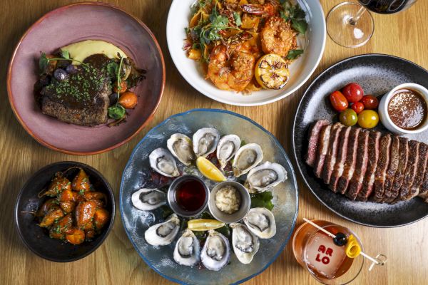 A table with diverse dishes: steak, pasta, oysters, roasted vegetables, ripe tomatoes, and a cocktail. Fine dining setting.