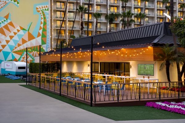An outdoor restaurant with string lights and colorful seating, adjacent to a hotel with balconies and a large geometric mural on a building side.