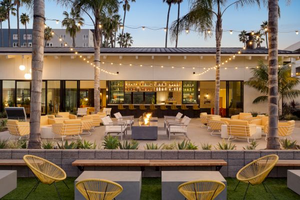 An outdoor seating area with yellow chairs, string lights, and palm trees. There's a bar at the back and a central fire pit surrounded by seating.