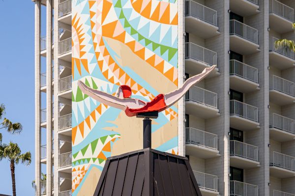 A building features a colorful geometric mural, with a rooftop sculpture of a person diving, set against a clear blue sky.