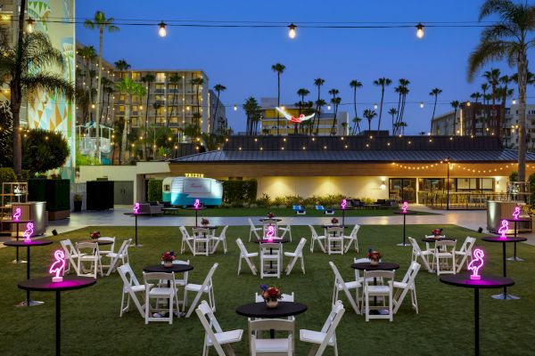 An outdoor venue with string lights, tables, chairs, neon signs, and palm trees at dusk.