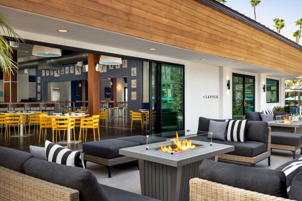 The image features an outdoor seating area with a fire pit, surrounded by modern wicker furniture. There's an indoor dining space with yellow chairs.