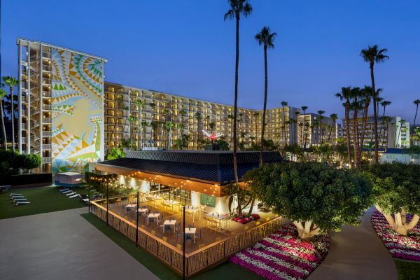 The image shows an outdoor dining area with lights, surrounded by trees and flowers, next to a tall hotel building with a mural and palm trees.