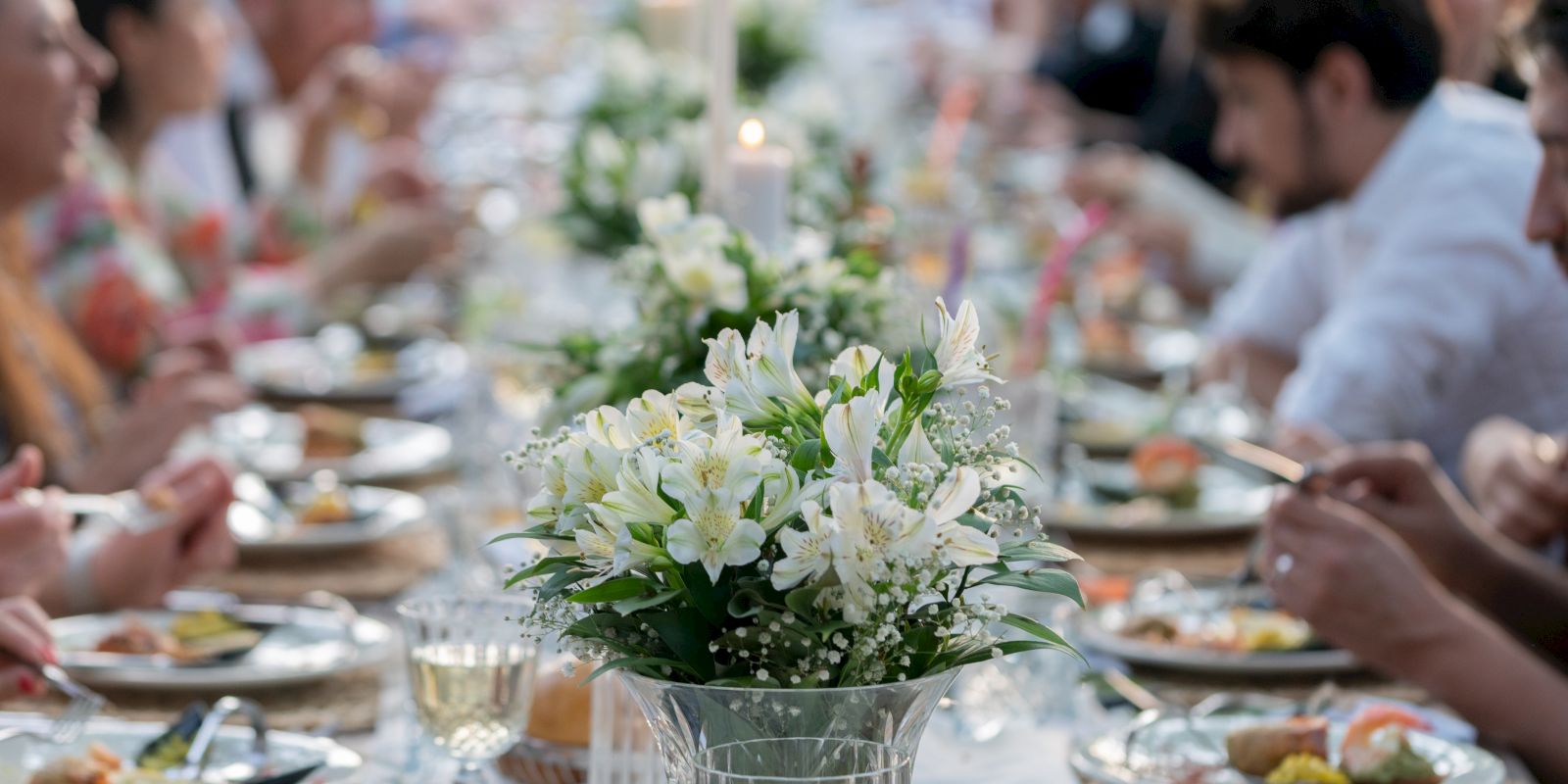 People are dining at a long table adorned with white flowers, enjoying a meal together in a communal setting outdoors.