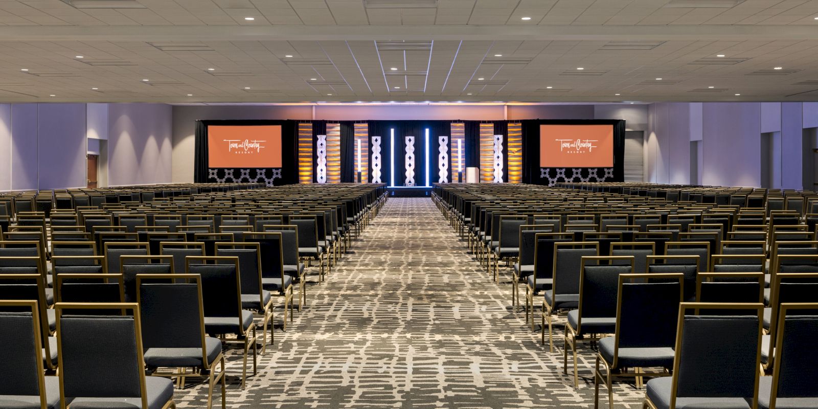 A large conference room with rows of chairs facing a stage with two screens displaying text and graphics, set up for a presentation or event.