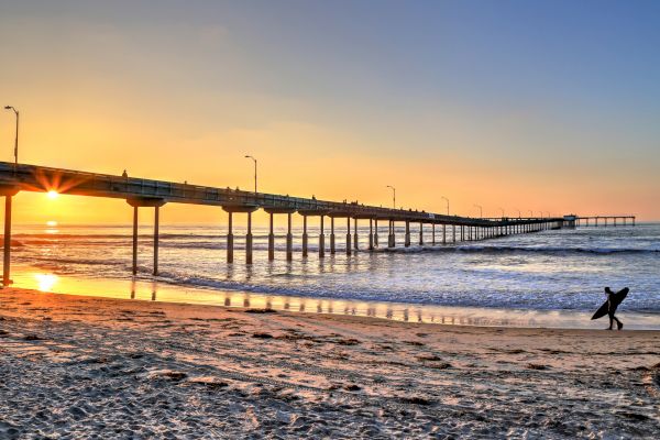 A serene beach scene at sunset, with a pier extending into the ocean and a lone surfer walking along the shore.