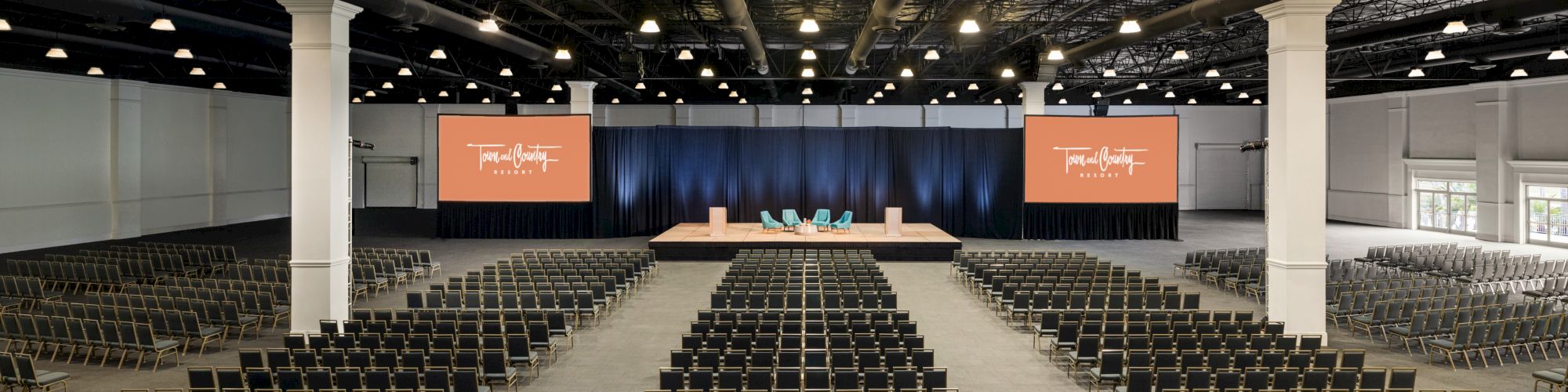 The image shows a large, empty conference room with rows of chairs facing a stage with a podium and screens on each side, illuminated by ceiling lights.