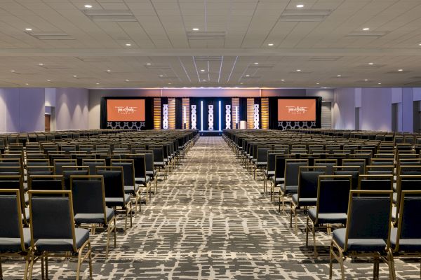 A spacious conference room with numerous rows of empty chairs facing a stage with large screens and modern lighting.