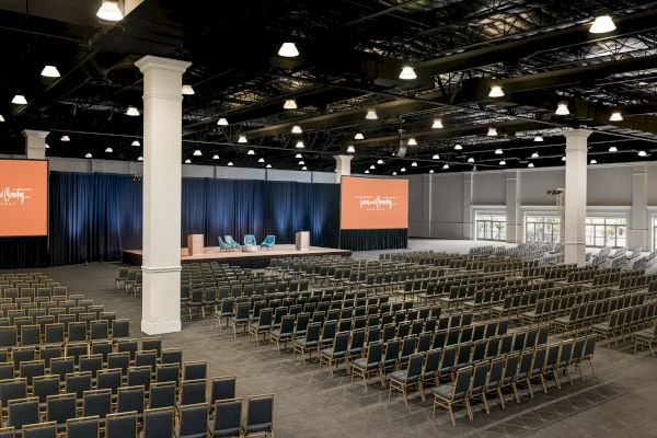 A large conference room with rows of chairs, a stage with chairs, and two large screens displaying text on either side of the stage.