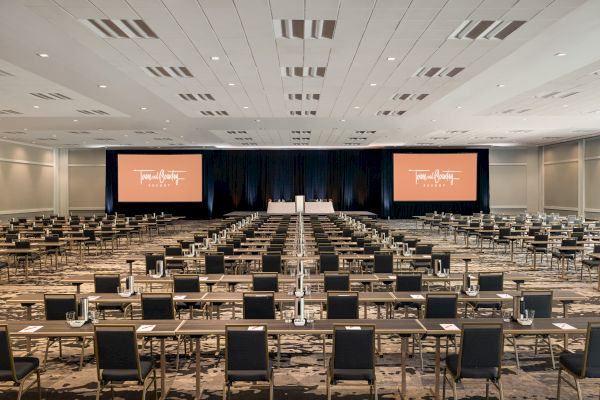 A large conference room with many rows of tables and chairs facing a stage with two large screens displaying a presentation.