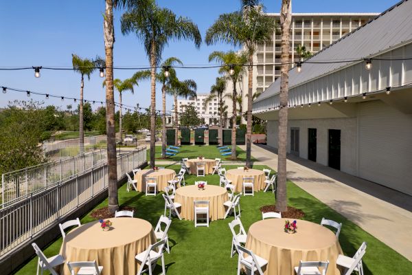 An outdoor event setup with round tables covered in beige tablecloths, white chairs, string lights, palm trees, and buildings in the background.