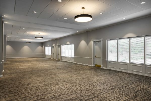 The image shows a large, empty room with carpeted flooring, multiple windows, and ceiling lights, creating a bright and open space.