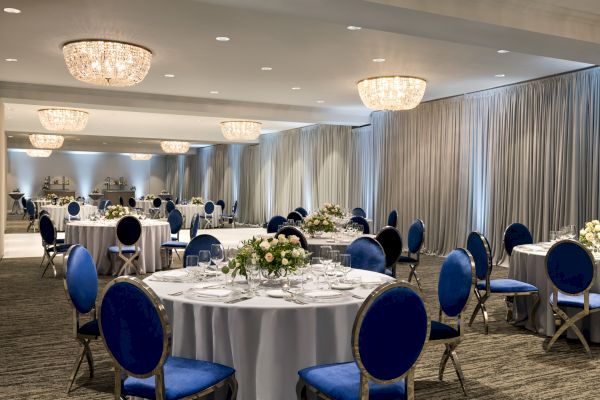 Elegant banquet hall setup with round tables, blue chairs, floral centerpieces, chandeliers, and draped curtains.