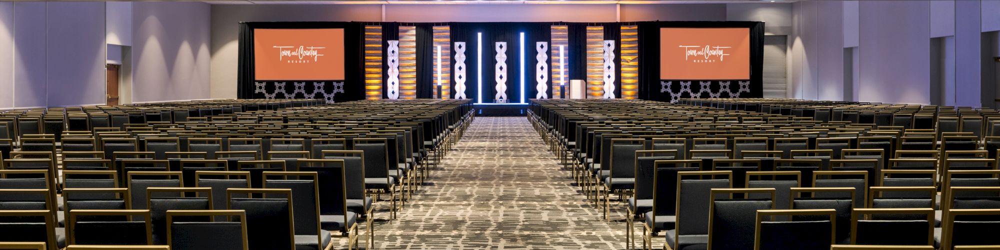 A large conference room with rows of chairs facing a stage with two screens on either side, featuring a patterned carpet and overhead lighting.