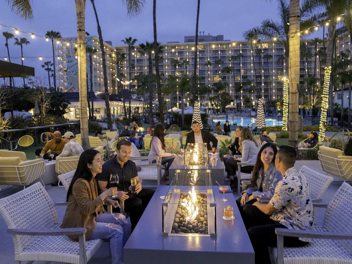 People are sitting around a fire pit at an outdoor venue with string lights and palm trees, chatting and enjoying drinks in the evening.