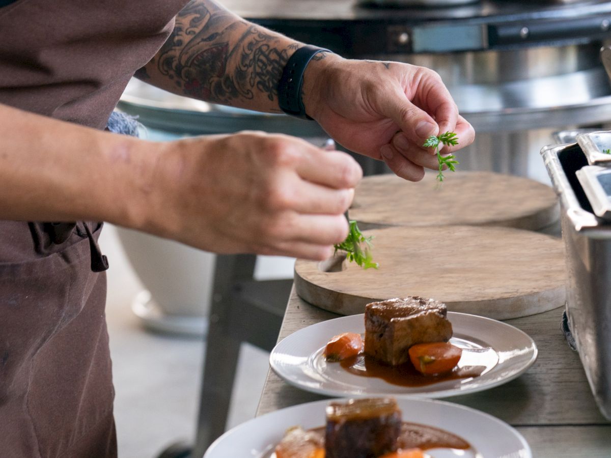 A person is garnishing plated meat dishes in a kitchen setting, standing next to cooking equipment and wearing a brown apron with tattoos on their arm.