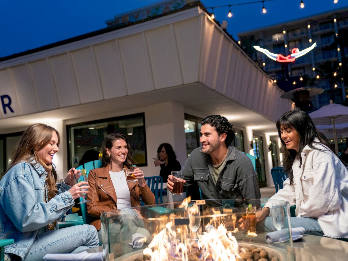 A group of four people sitting around a fire pit, smiling and enjoying drinks at an outdoor venue during the evening.