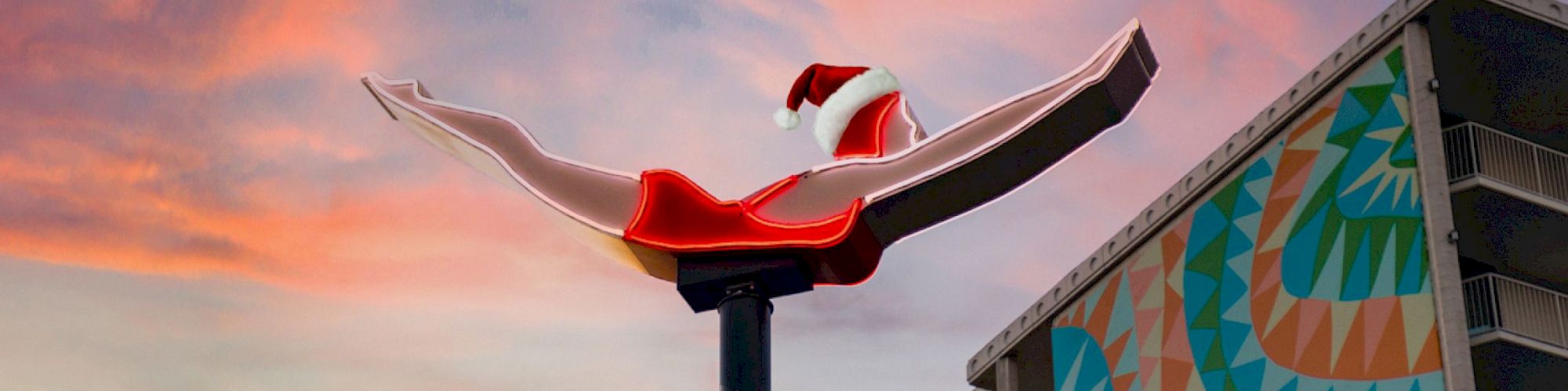 A neon sign of a diver wearing a Santa hat is displayed against a colorful sunset sky near a building with a geometric mural.