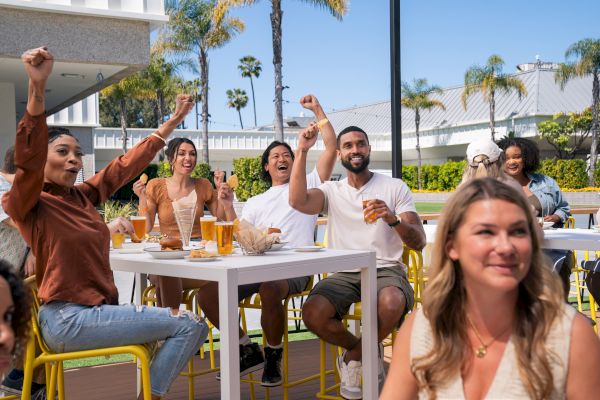 A group of people are seated outdoors at a table with drinks and snacks, cheering and celebrating. There is palm tree scenery in the background.