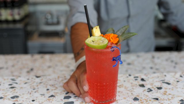 A person is holding a red cocktail garnished with lime, pineapple, and an orange flower, with a straw and a blue stirrer on a speckled countertop.