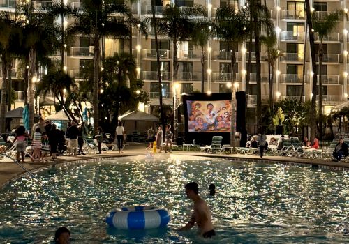 The image shows a nighttime scene with people swimming in a pool while a movie plays on a screen beside the pool, surrounded by lit buildings.