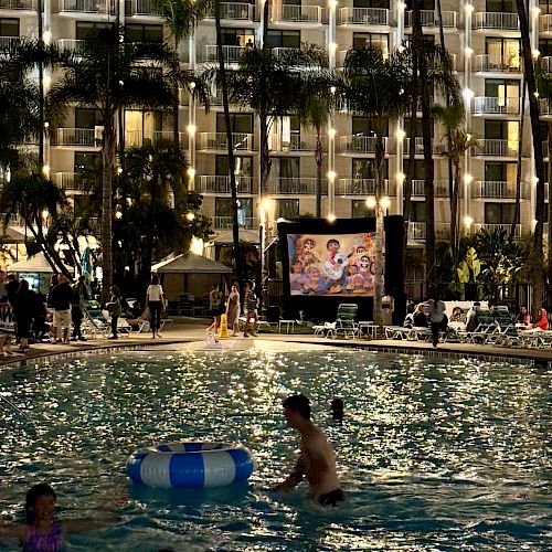 The image shows a nighttime scene with people swimming in a pool while a movie plays on a screen beside the pool, surrounded by lit buildings.