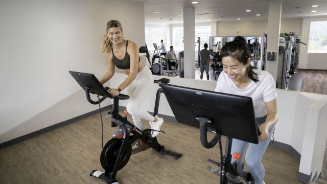 Two women are using exercise bikes with screens in a modern gym, smiling as they workout.