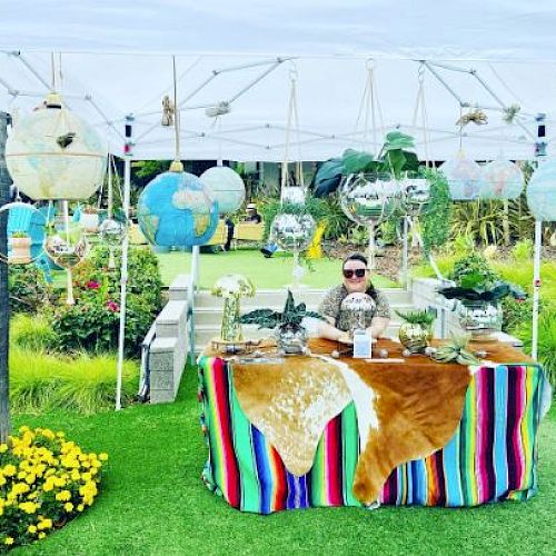 A person sitting behind a table with a colorful cloth, surrounded by hanging plants and terrariums at an outdoor market.