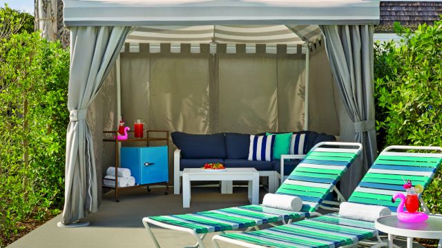 A poolside cabana with striped lounge chairs, a small table with a pink flamingo float, and a shaded area with a sofa and storage cabinet.