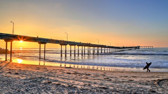 A scenic beach at sunset with a long pier extending into the ocean and a surfer walking along the shore carrying a surfboard.