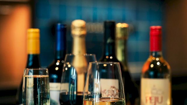 The image shows a bar setup with various bottles of wine and three filled wine glasses placed on a wooden counter.