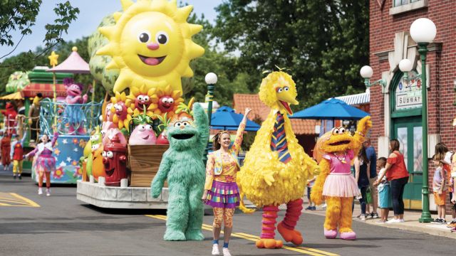 A colorful parade with people in costumes, including a large bird, walking down a street. Bright float with a smiling sun and other characters behind them.