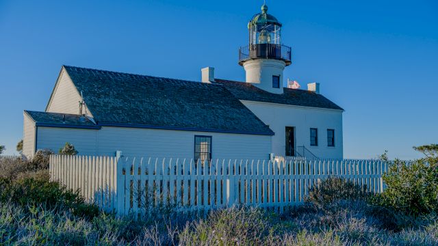 The image shows a white lighthouse with a green roof and a white picket fence around it, set against a clear blue sky.