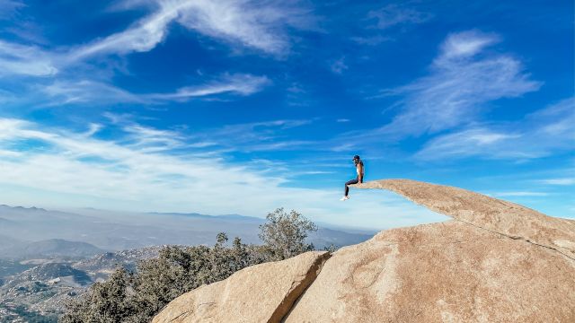 A person sits on a protruding rock edge high above a scenic landscape with a vast blue sky and scattered clouds in the background.