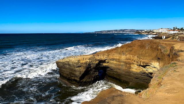 A coastal landscape with rocky cliffs, waves crashing against them, and clear blue skies above, extending towards the horizon.