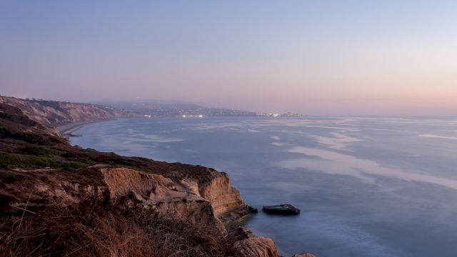 The image captures a serene coastal view at dusk with cliffs, calm water, and a distant shoreline with scattered lights, under a clear sky.