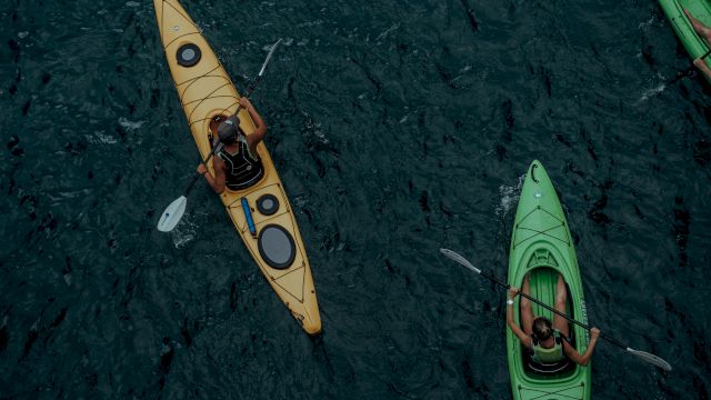 The image shows two people kayaking in dark water, one in a yellow kayak and the other in a green kayak, paddling alongside each other.