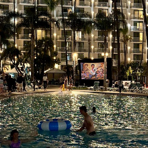 People are enjoying a nighttime swim in a pool with a large building and an outdoor movie screen in the background.