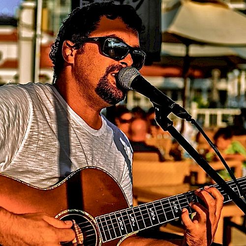 A man wearing sunglasses is singing and playing an acoustic guitar into a microphone at an outdoor setting with people in the background.