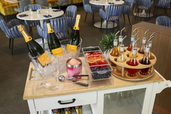 A bar cart with champagne bottles, glasses, fruit garnishes, and assorted syrups on a wooden counter in a restaurant setting.