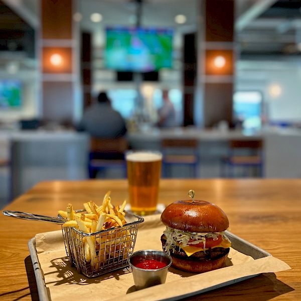 A burger, fries, and a glass of beer on a table in a restaurant; background shows patrons seated at a bar with televisions on the wall.