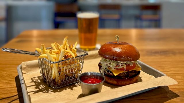 A burger, fries, and a glass of beer on a table in a restaurant; background shows patrons seated at a bar with televisions on the wall.