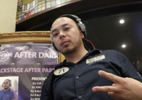 A DJ wearing headphones and a black shirt stands behind a turntable and mixer, making a peace sign with his fingers. There's a poster in the background.