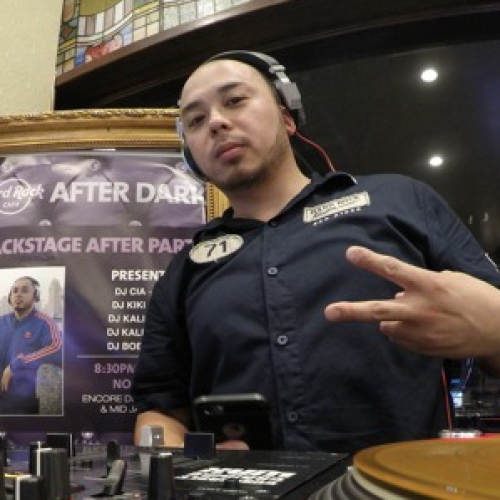 A DJ wearing headphones and a black shirt stands behind a turntable and mixer, making a peace sign with his fingers. There's a poster in the background.