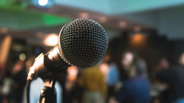 The image features a close-up of a microphone with a blurred background, possibly at an event or gathering with people mingling.