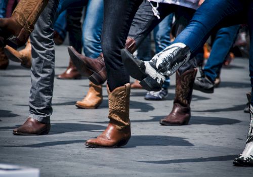 People wearing cowboy boots in various colors and designs are dancing on a street or an open space, lifting their legs in unison.