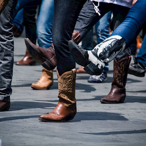 People wearing cowboy boots in various colors and designs are dancing on a street or an open space, lifting their legs in unison.