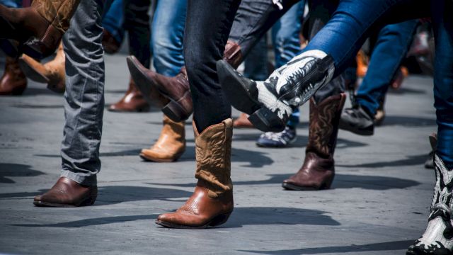 People wearing cowboy boots in various colors and designs are dancing on a street or an open space, lifting their legs in unison.