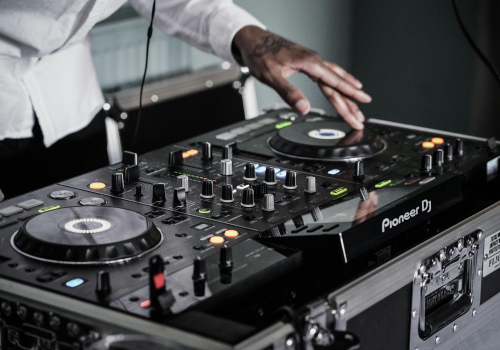 A DJ is using a Pioneer mixer and turntables, adjusting knobs and discs with a hand, wearing a white shirt.